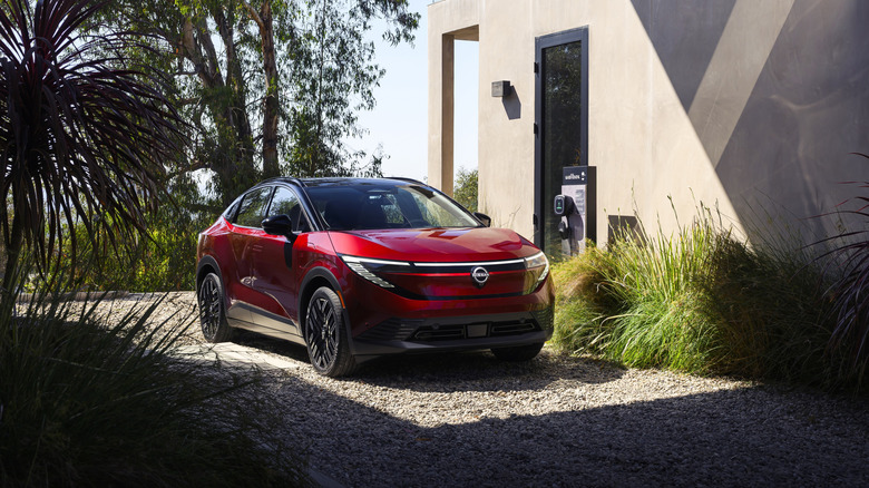 Pogled od spredaj 3/4 rdečega nissana leafa 2026, parkiranega na dovozu poleg polnilnice za električna vozila