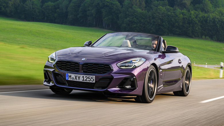 Temno vijoličen kovinski BMW Z4 se vozi s spuščeno streho po podeželski cesti z zeleno travo in drevesi v ozadju.