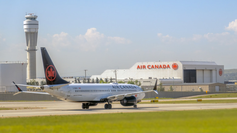 Letalo Boeing 737 MAX 8 Air Canada taksira na vzletno-pristajalni stezi s kontrolnim stolpom in hangarjem Air Canada v ozadju.