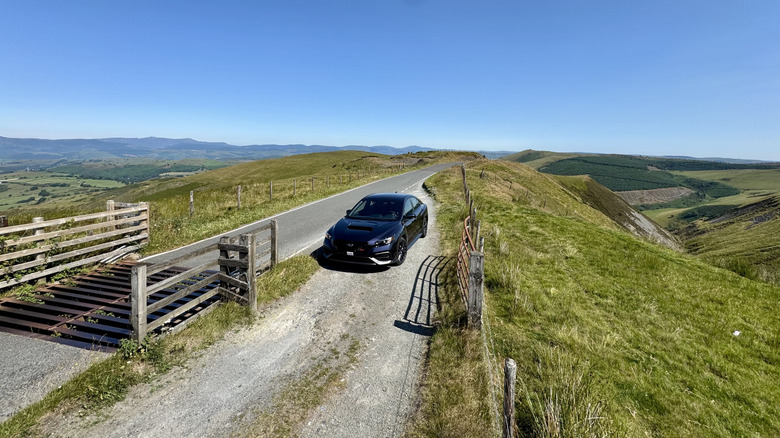 Posnetek od zgoraj vozila Galaxy Purple Pearl WRX, parkiranega na makadamskem križišču poleg lovilca krav na vrhu travnate gore s pogledom na travnike in modro nebo