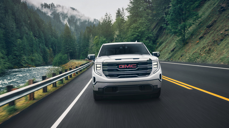 Pogled od spredaj na belo GMC Sierra 1500 2024, ki se vozi skozi gorski gozd ob reki