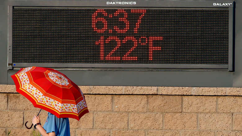 Moški z dežnikom na sončen dan hodi mimo znaka, ki prikazuje temperaturo 122ºF