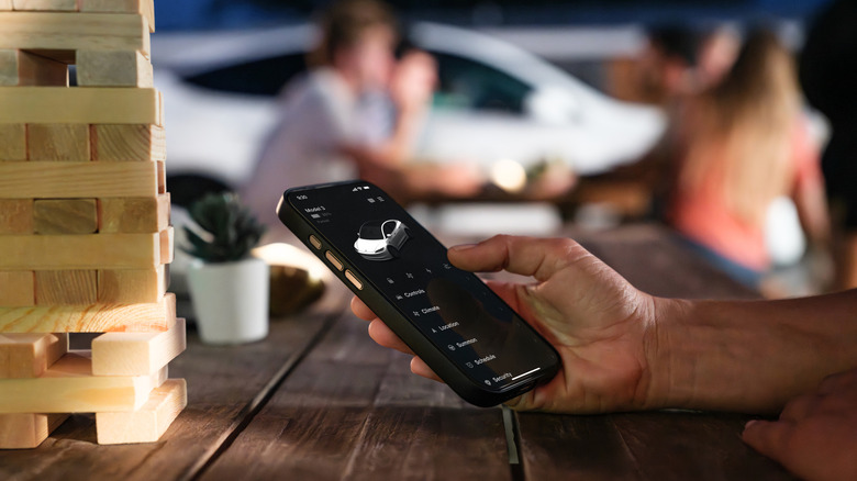 Roka drži telefon z aplikacijo Tesla na zaslonu, na mizi z igro Jenga na levi in ​​ljudmi v ozadju