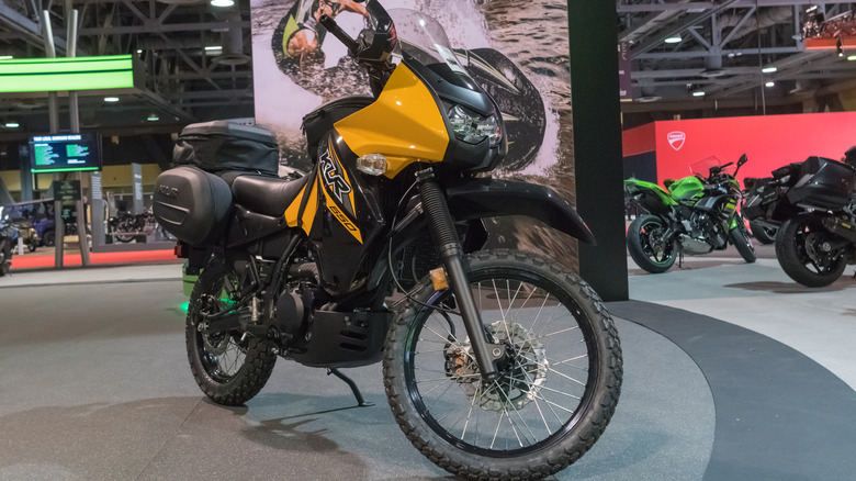 Novi črno-rumeni Kawasaki KLR 650 na razstavnem prostoru z različnimi drugimi motocikli v ozadju