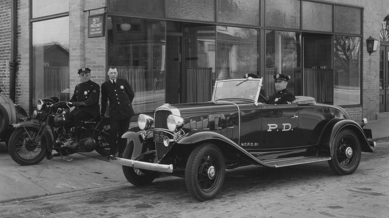 Črno-bela fotografija policistov v policijskem avtomobilu in na motorju pred policijsko postajo leta 1929.
