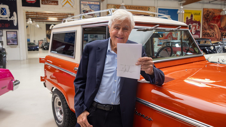 Jay Leno stoji s komolcem na oranžnem Broncu v svoji garaži in drži podpisan račun