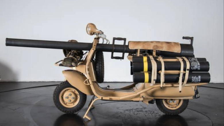 Vespa 150 TAP peščene barve, opremljena s pištolo, v studiu z belo steno in črno okroglo mizo.