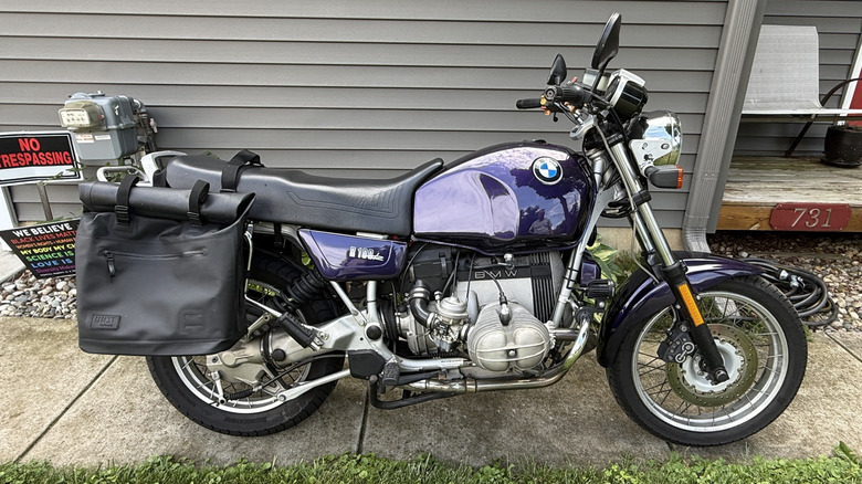Vijolični BMW R100R letnik 1992, parkiran na pločniku pred sivo hišo.