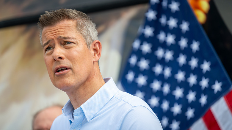 Ameriški minister za promet Sean Duffy je videti zmeden, medtem ko nekje stoji pred ameriško zastavo med govorom.